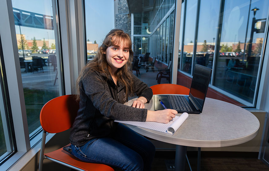 Student sitting at a table working on computer, smiling at camera