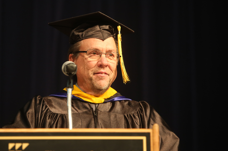 Image of a past Distinguished Alumnus standing at a podium giving a speech for graduation, wearing a cap and gown.