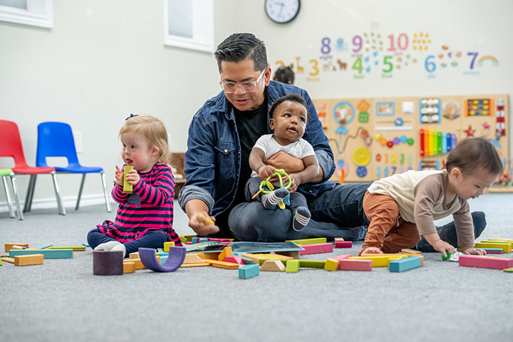 Three small toddlers sit in closely with a male childcare worker, as they learn through play together.