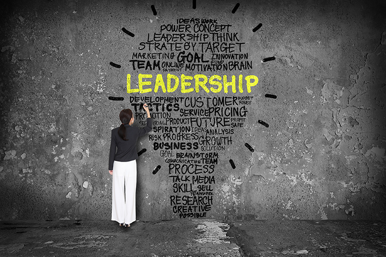 Woman standing at gray wall with words defining leadership in the shape of a lightbulb and 'leadership' in large bright yellow